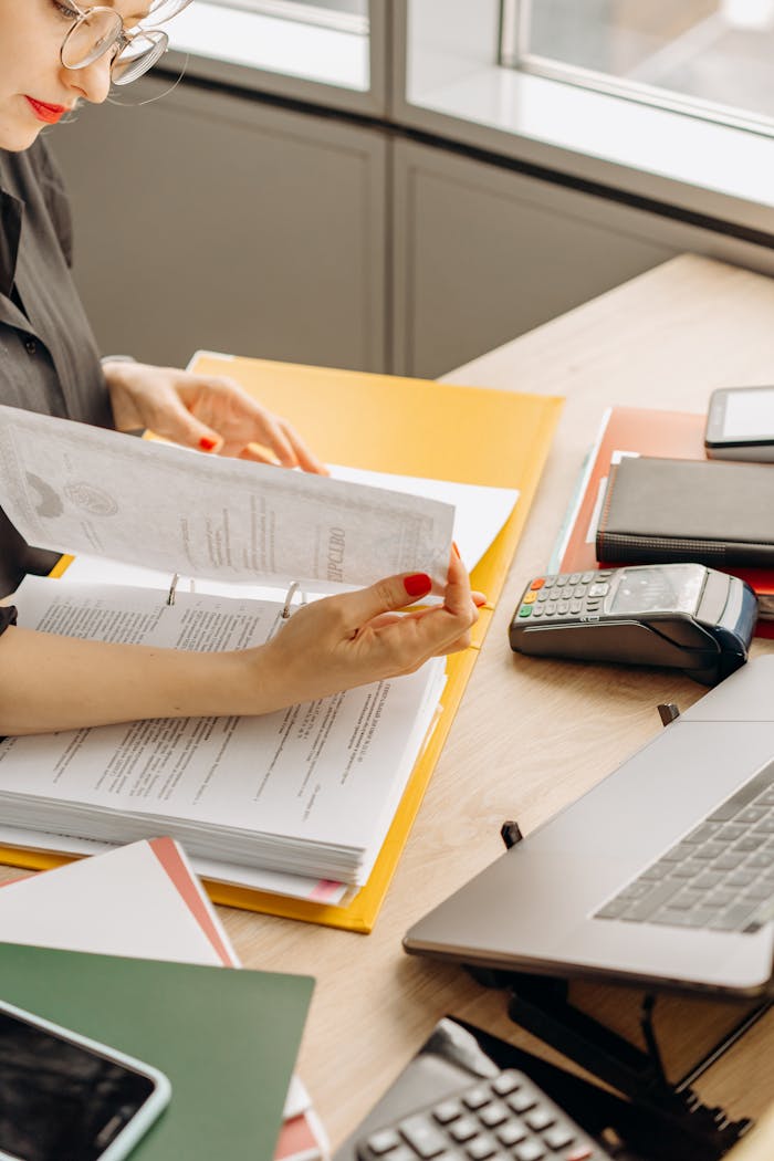 A woman flipping through financial documents on a wooden desk with various office supplies.